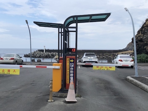 海濱停車場出入口實景：車道上方有金屬與玻璃雨遮及門架，淨高明顯低於約一層樓，高頂露營車無法安全通過；提醒租車新手留意路邊老宅雨遮、停車場與巷口低矮構造物，寧可繞道或改停露天車位也不要硬開