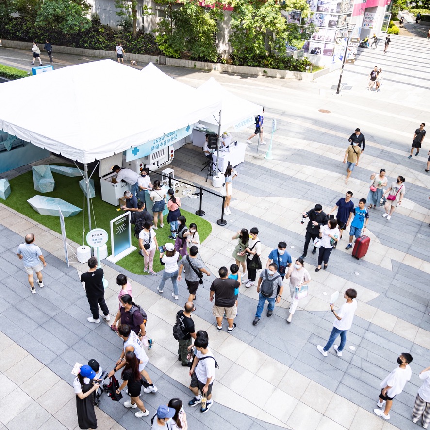 空拍視角：戶外廣場上 SNOW TOUCH 急救站白色棚區、露營車與排隊動線，周邊行人與建物呈現都市快閃規模感