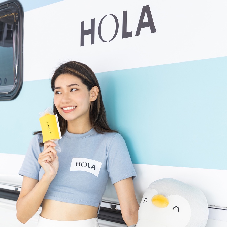 模特身穿胸前 HOLA 字樣上衣，倚靠白藍配色露營車車身，車側有 HOLA 大字標誌，手邊有企鵝玩偶一角，明亮戶外形象照