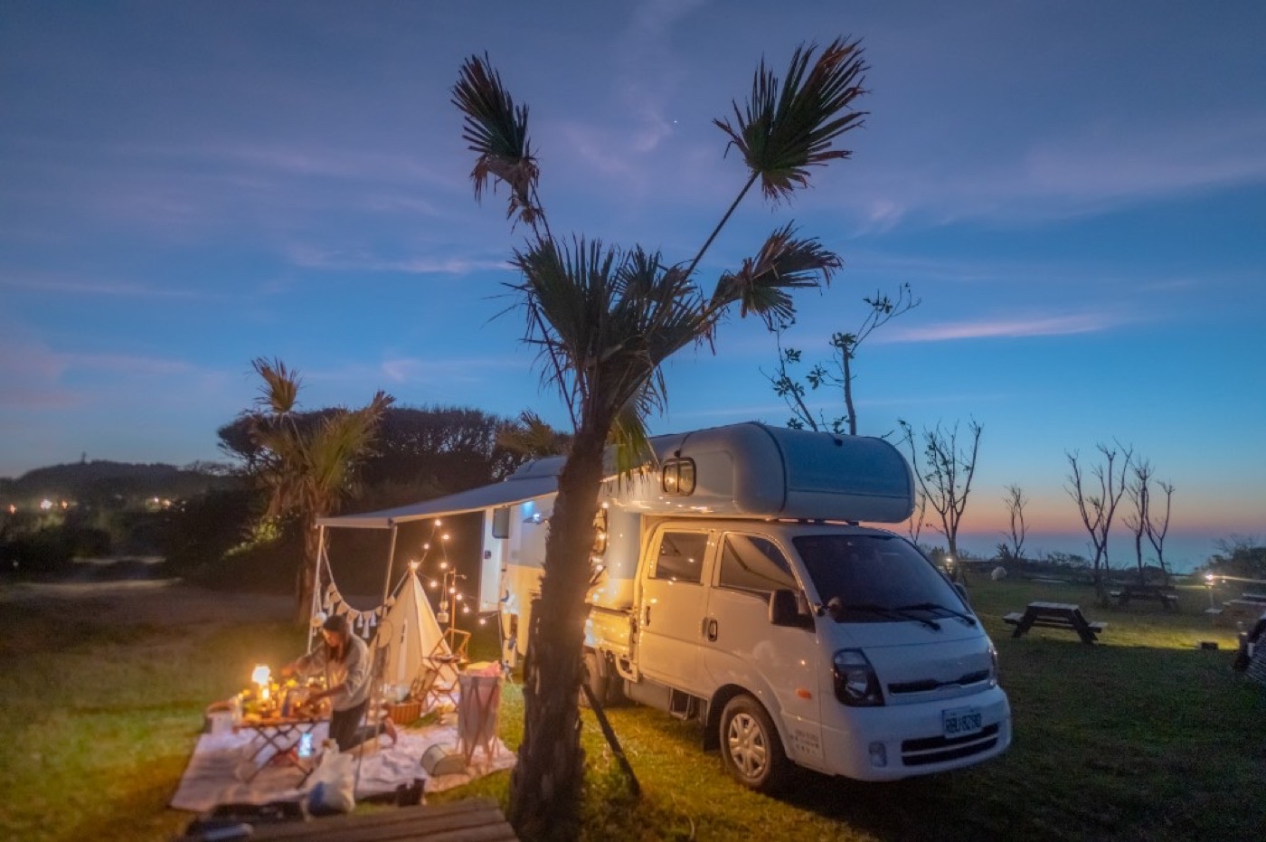 White campervan on Bunker Campsite grass at blue hour, string lights, folding wood table and small teepee—romantic van-side dinner setup