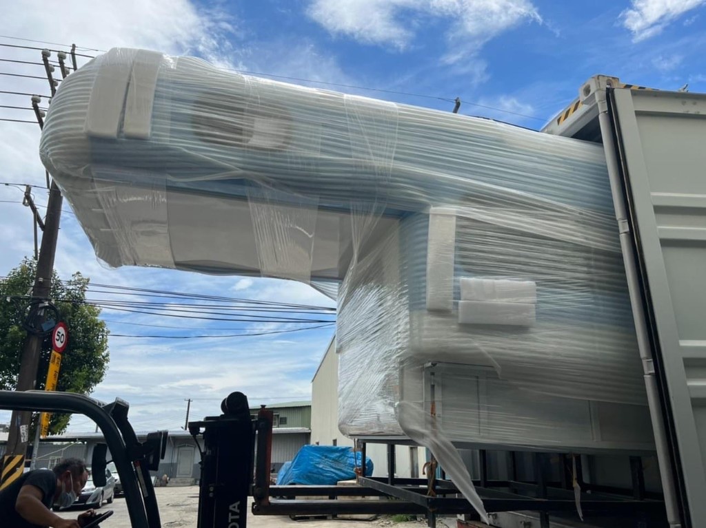 Team with forklift unloading new white campervan shell wrapped in protective film from container, cab-over shape visible