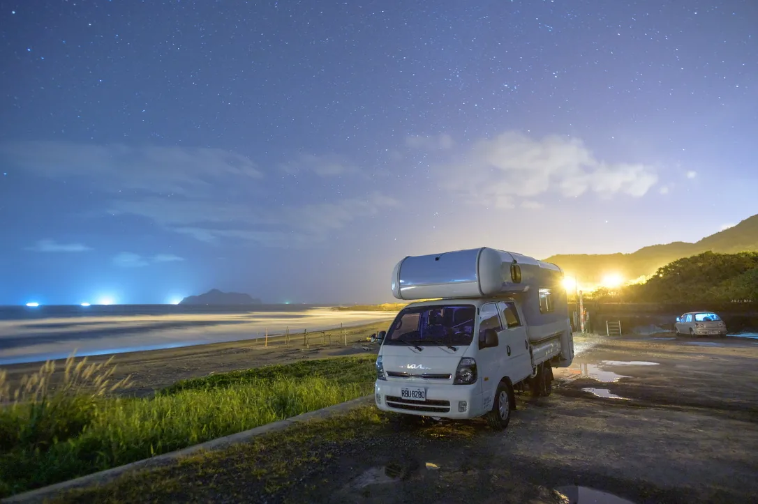 星空下露營車停在海岸旁，遠方是被月光照亮的沙灘與海面