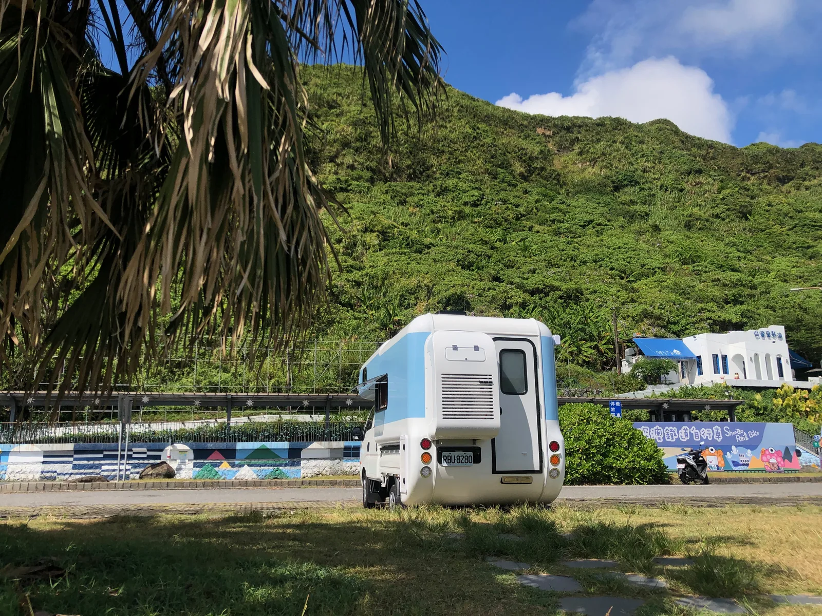 藍白露營車停在大片翠綠山坡前，棕櫚葉與藍天形成臺灣亞熱帶山景