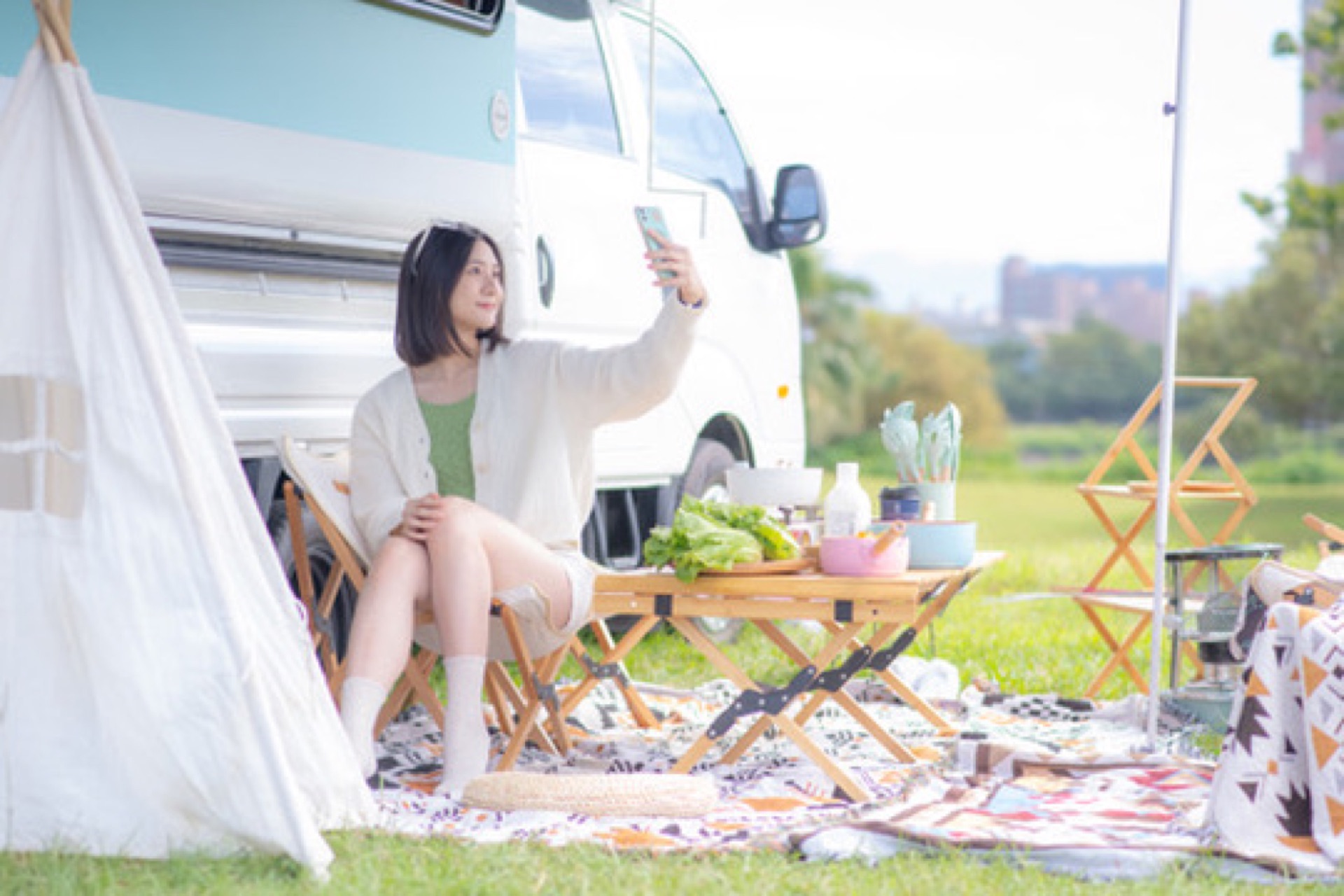 短髮女生坐在露營車旁摺疊椅上拿手機自拍微笑，木桌、圖騰野餐墊與小帳篷布置，遠方天際線模糊，呈現 aesthetic 風格露營車輕旅行與自由自在氛圍