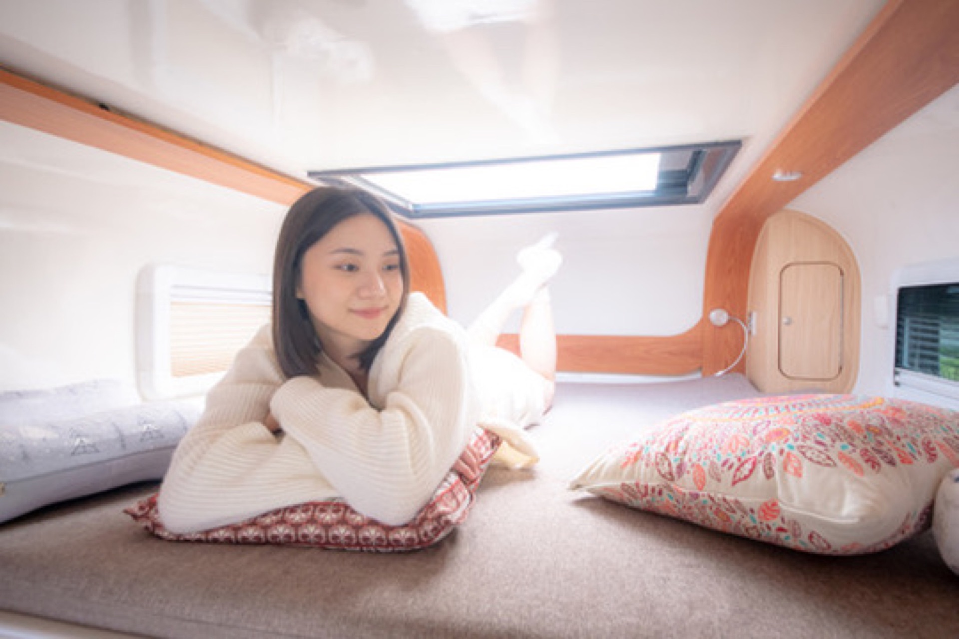 年輕女性在露營車上層額頭床俯臥休憩，木質線條與長方形天窗透入柔和日光，多色抱枕與閱讀燈，呈現睡醒後慵懶舒適的露營車寢區與慢旅體驗