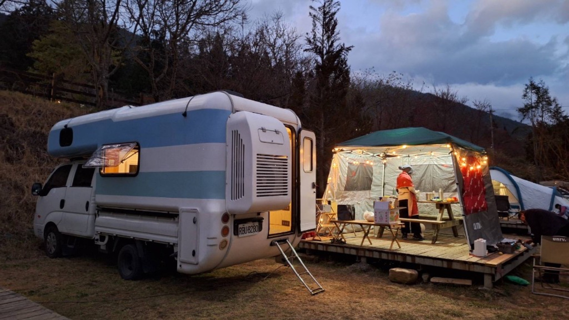 Dusk white-and-blue campervan beside grass pitch, wooden deck and Edison string lights on net shelter, travelers preparing food, warm light from doors and windows—relaxed highland campervan setup