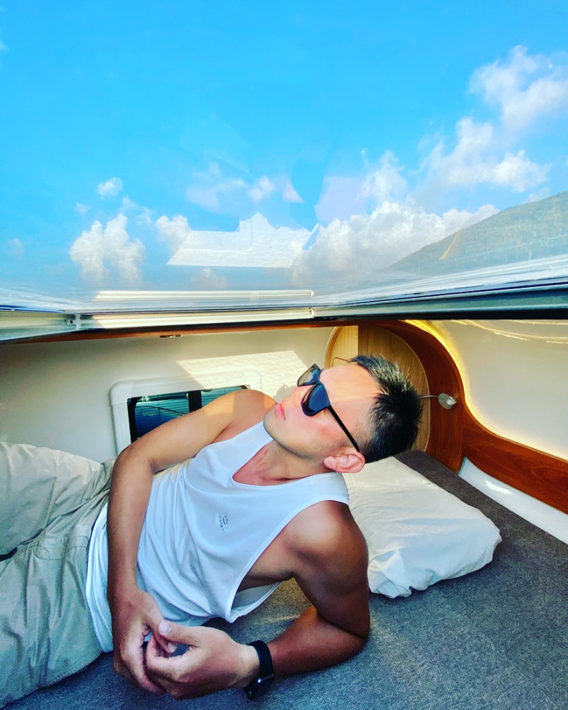 Traveler on campervan bed looking up through skylight at blue sky—relaxed post-swim AC break