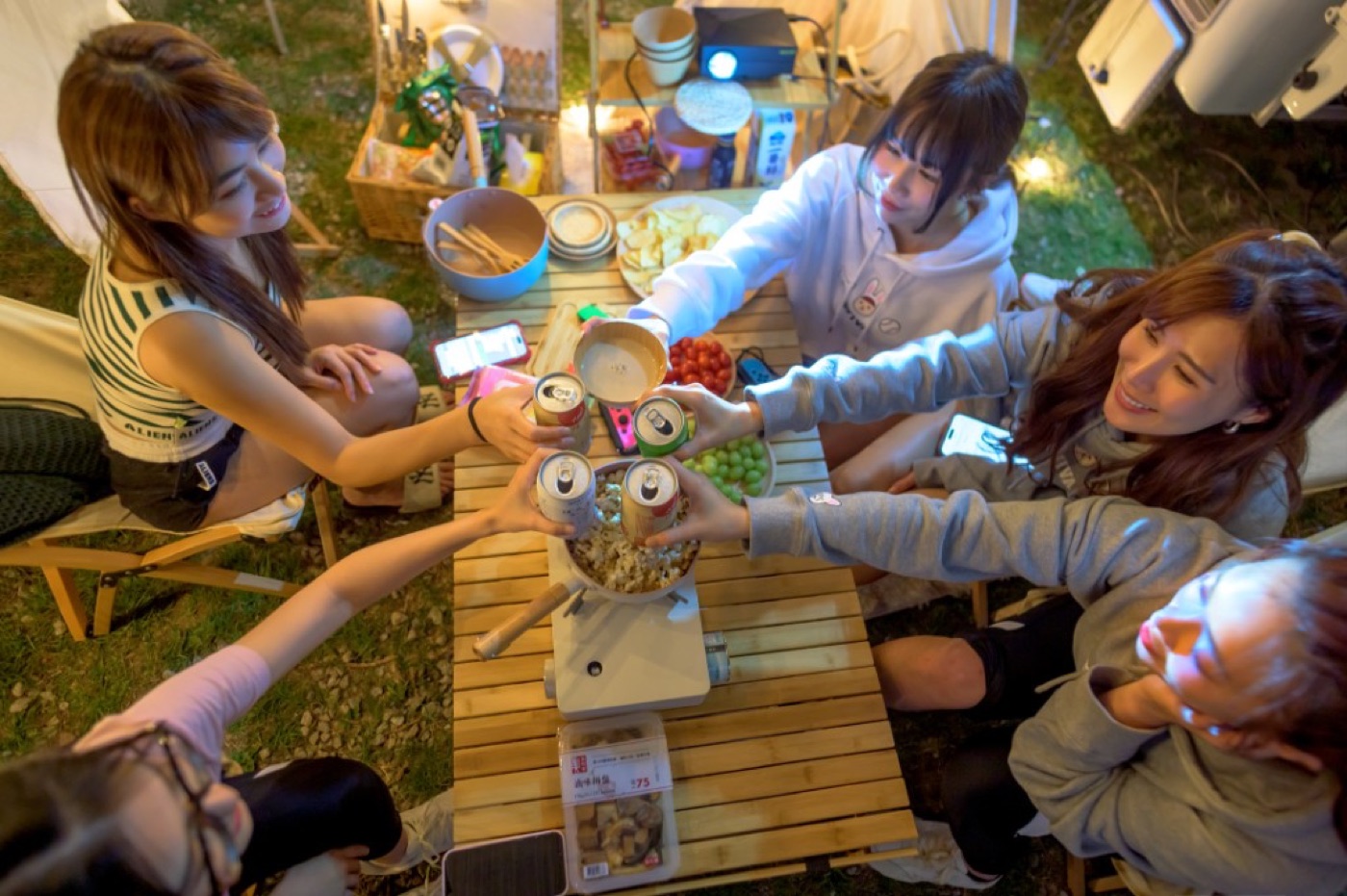 Overhead five women toasting cans at wood table, snacks fruit phones, camp lights night background