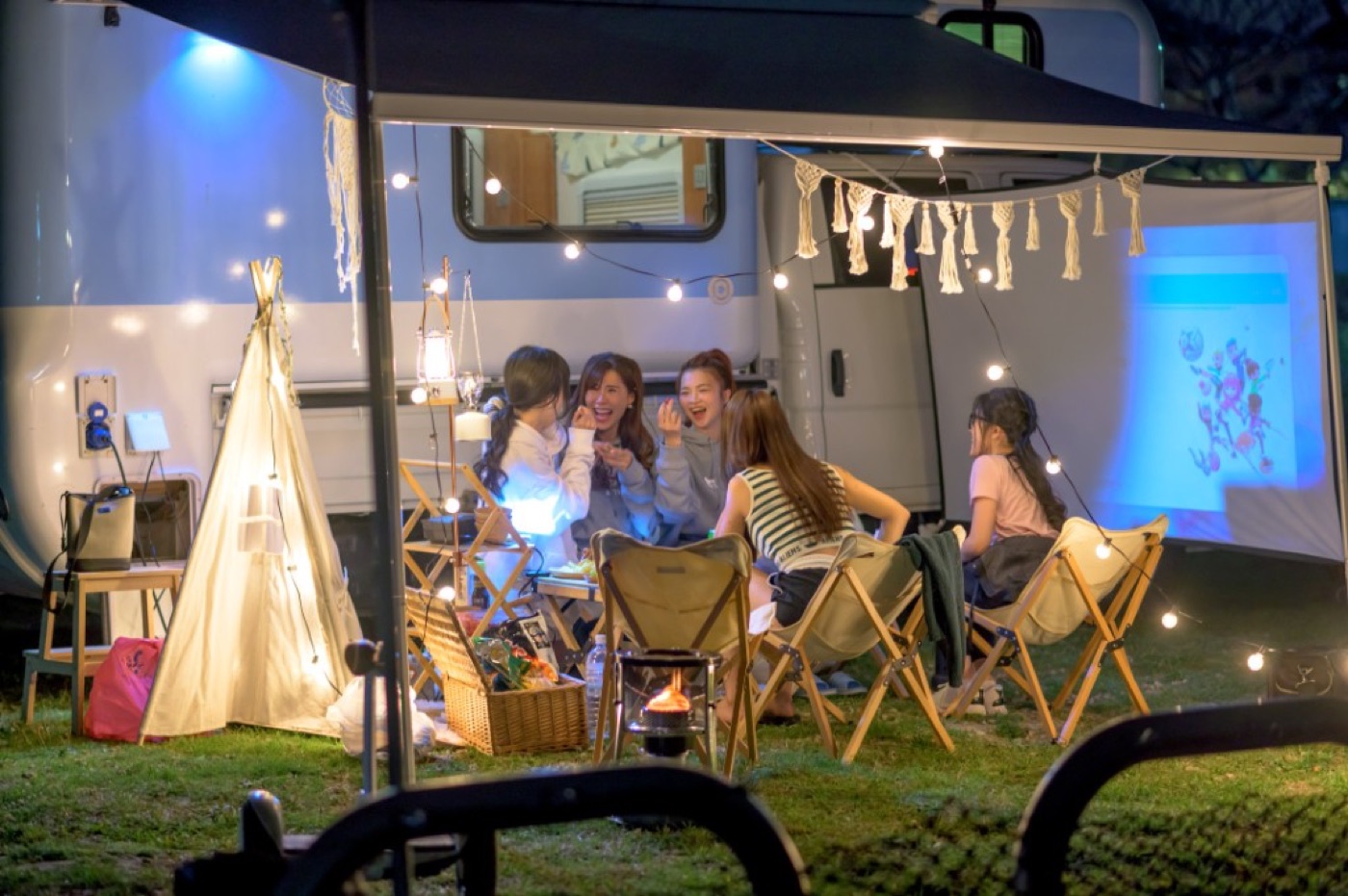 Night camp blue-white campervan side awning open, string lights small tent lighting five women chatting, projection and camp decor beside
