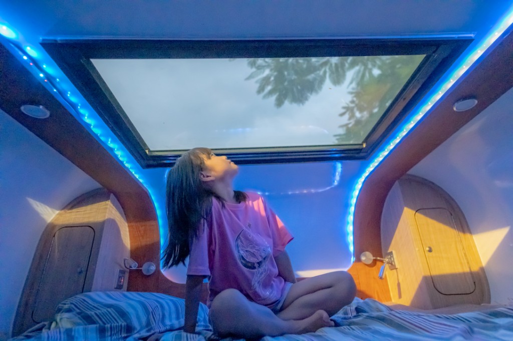 Child on the cab-over bed looking up through the skylight at night trees, blue mood lighting and wood interior